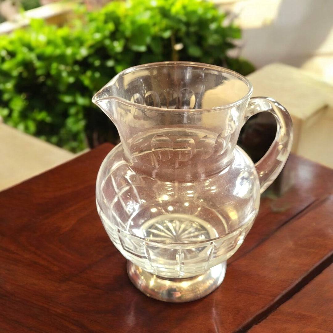 Jarra de cristal tallado con base de metal plateado sobre mesa de madera, ideal para decoración elegante en interiores o exteriores.