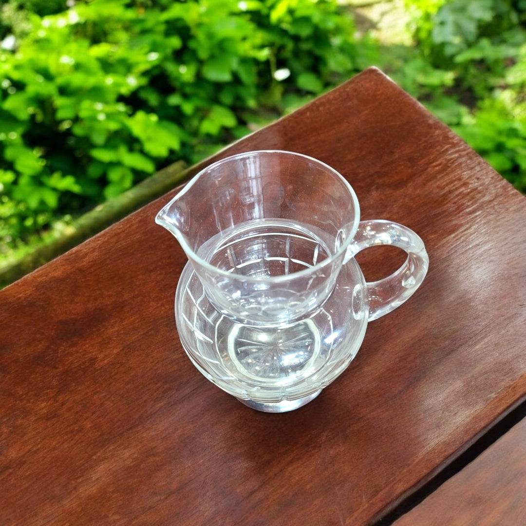 Jarra de cristal tallado con base de metal plateado sobre mesa de madera, ideal para decoración elegante en interiores o exteriores.