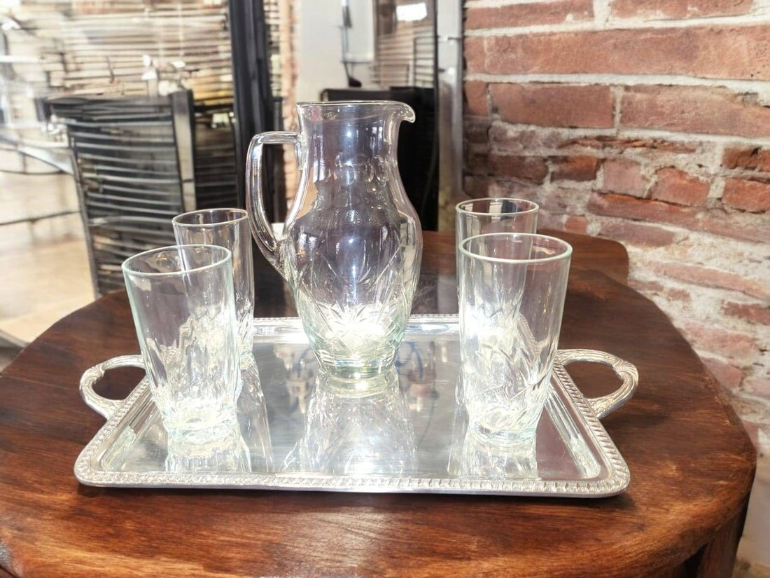 Juego de jarra de cristal y vasos tallados, elegante y clásico, sobre bandeja metálica decorativa en mesa de madera rústica. Ideal para decoración de cocina o comedor.
