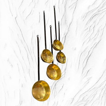 Lote de cazos antiguos con mango de forja y metal dorado colgados en pared blanca texturizada. Ideal para decoración rústica o vintage.