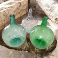  Tres damajuanas antiguas de cristal soplado en tonos verde y transparente, con un hermoso efecto martillado, reflejando la luz sobre una base de piedra.