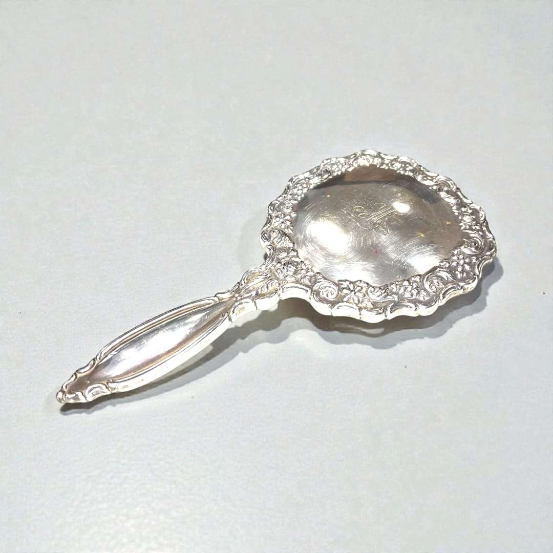 Este espejo de tocador destaca por su marco minuciosamente labrado, con curvas suaves y detalles cincelados que evidencian un trabajo artesanal refinado, propio de las piezas clásicas de tocador. 