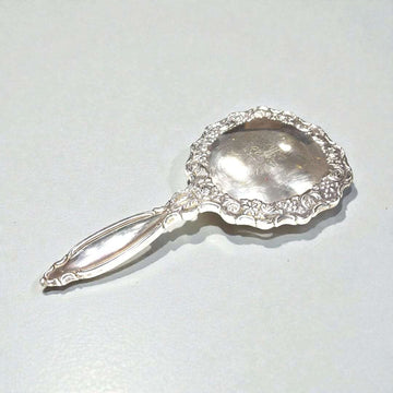 Este espejo de tocador destaca por su marco minuciosamente labrado, con curvas suaves y detalles cincelados que evidencian un trabajo artesanal refinado, propio de las piezas clásicas de tocador. 