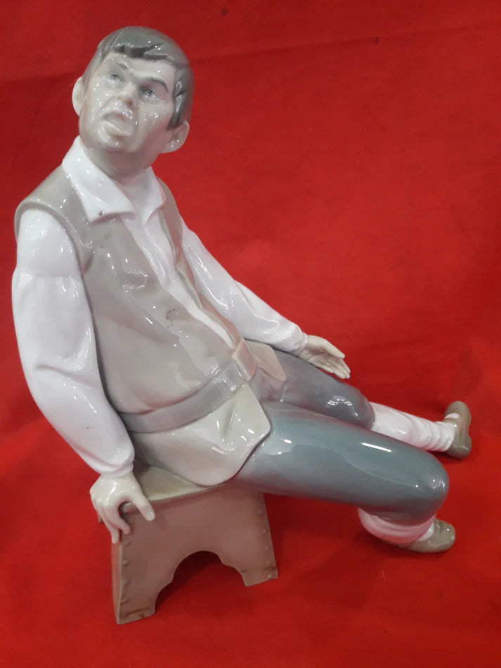 Figura Lladró de porcelana representando a un hombre sentado en un banco, con detalles en tonos beige, blanco y gris sobre fondo rojo.