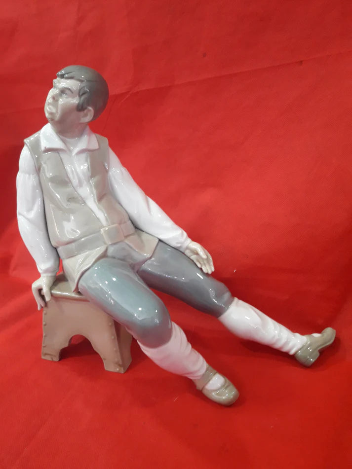 Figura Lladró de porcelana representando a un hombre sentado en un banco, con detalles en tonos beige, blanco y gris sobre fondo rojo.