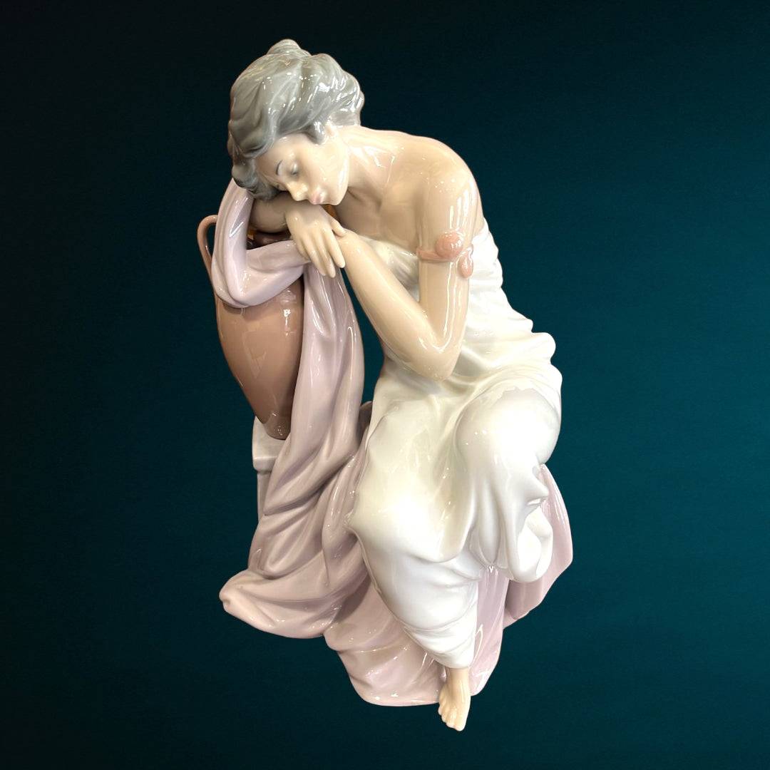 Figura Lladró 'Soñando Despierta' en porcelana fina, muestra a mujer recostada sobre ánfora con vestido blanco y rosa. Ideal para decoración elegante.