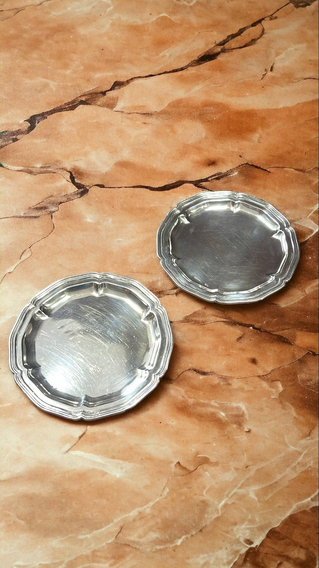 Platos decorativos de metal plateado con borde ondulado sobre superficie marmoleada. Añaden elegancia clásica a la decoración de mesa.