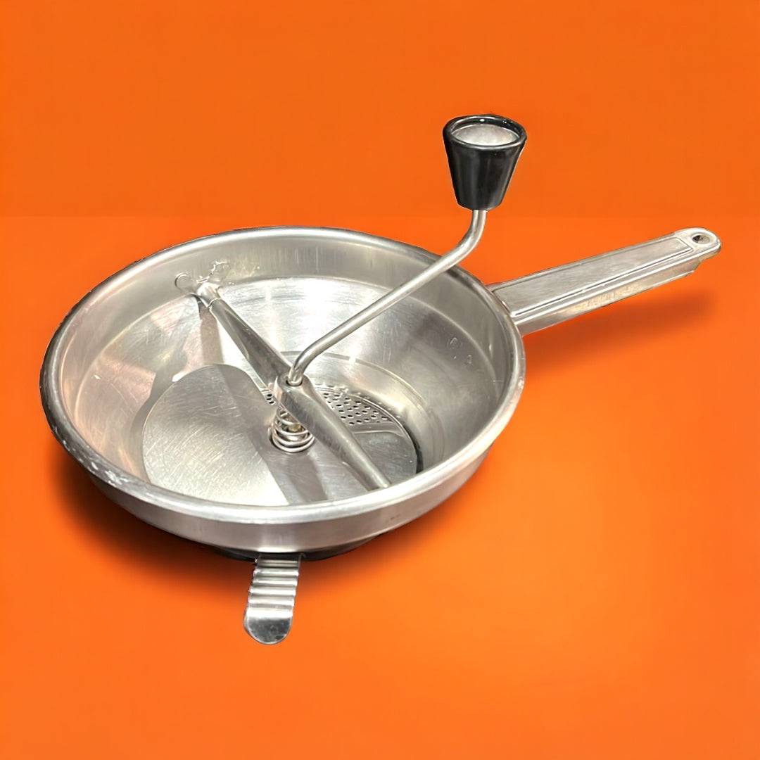 Pasapurés antiguo de aluminio con manivela negra sobre fondo naranja. Ideal para decoración vintage en cocinas retro o rústicas.