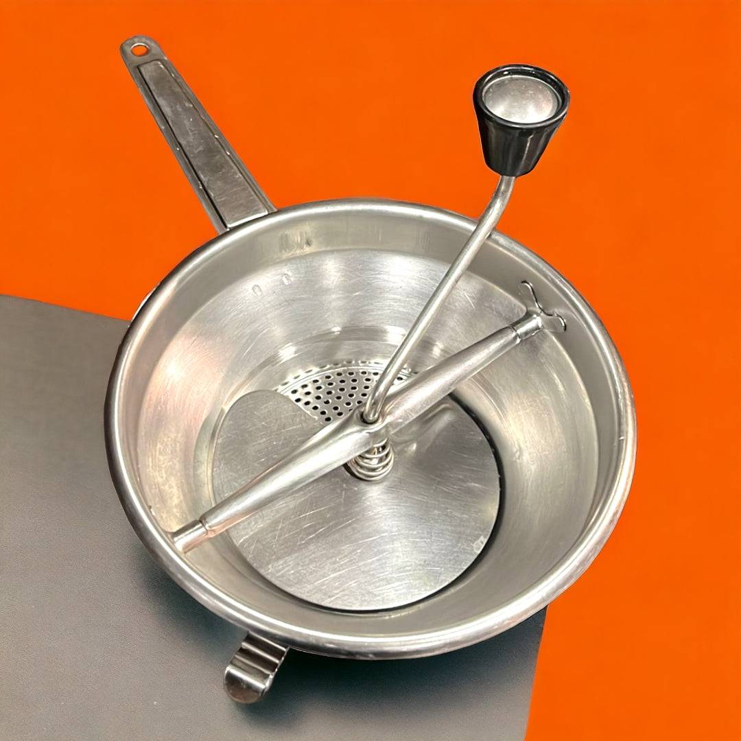 Pasapurés antiguo de aluminio con manivela negra sobre fondo naranja. Ideal para decoración vintage en cocinas retro o rústicas.