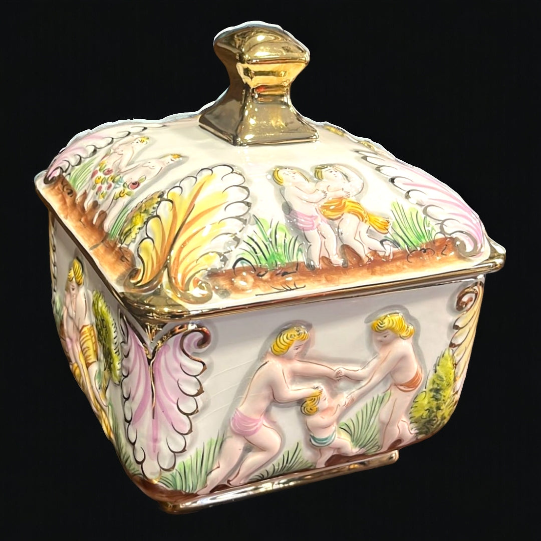 Bombonera de porcelana decorada con detalles dorados y escenas clásicas en relieve. Ideal para decorar y almacenar dulces. Medidas 17x17xH21 cm.