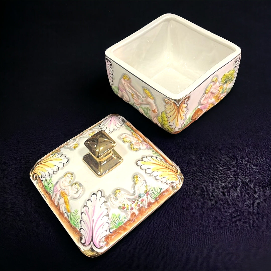 Bombonera de porcelana decorada con detalles dorados y escenas clásicas en relieve. Ideal para decorar y almacenar dulces. Medidas 17x17xH21 cm.