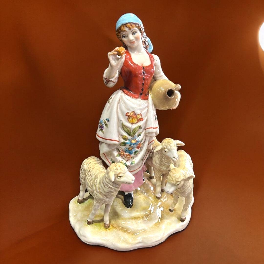 Figura Señora con Ovejas en porcelana exclusiva, con vestido tradicional, cántaro y tres ovejas sobre base decorativa. Ideal para decoración elegante.