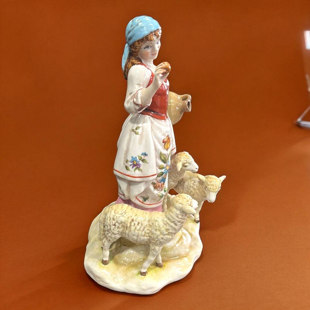 Figura Señora con Ovejas en porcelana exclusiva, con vestido tradicional, cántaro y tres ovejas sobre base decorativa. Ideal para decoración elegante.