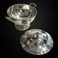 Sopera clásica de metal plateado, elegante y decorativa, con tapa desmontada y asas laterales, ideal para decoración de mesa sofisticada.