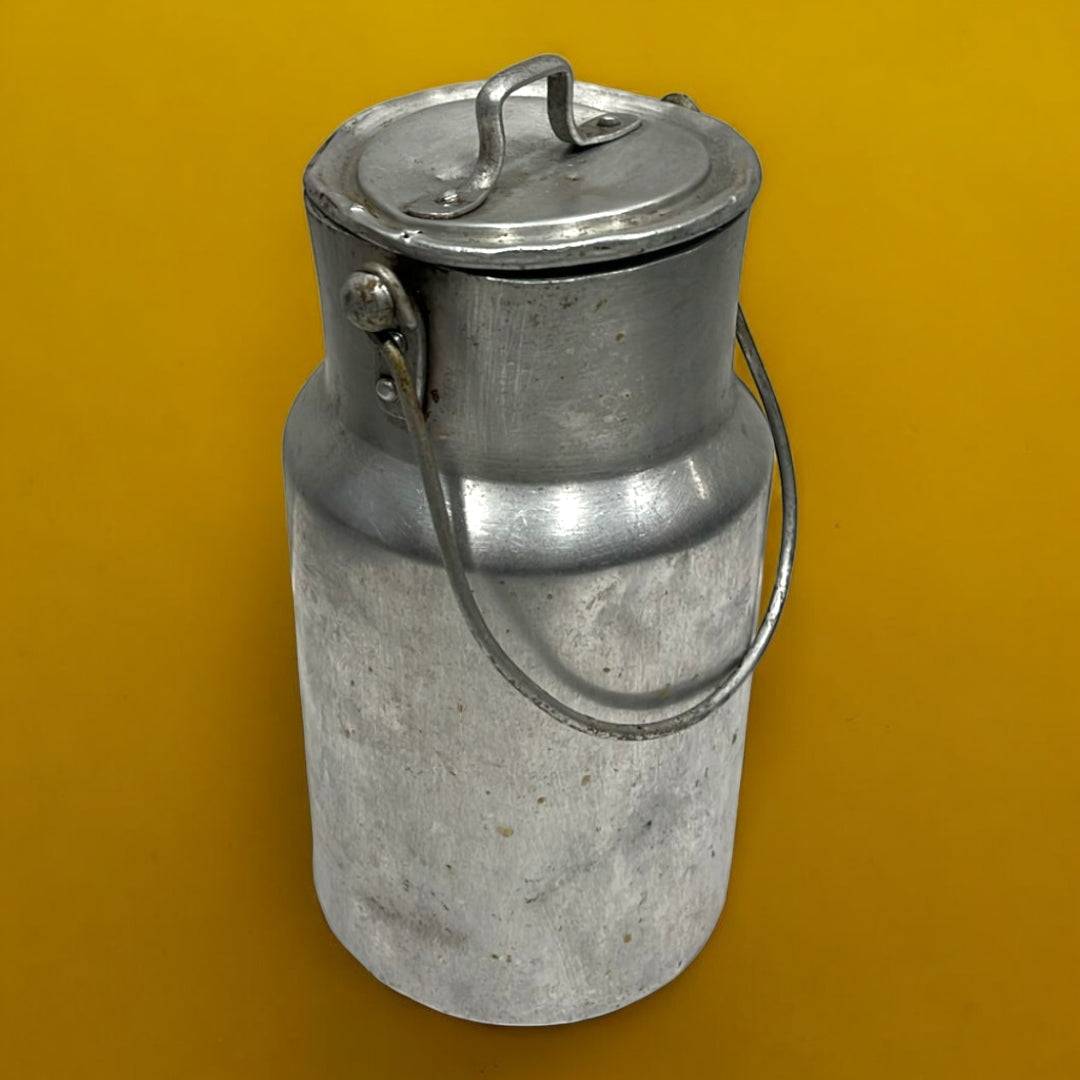 Lechera antigua de aluminio con tapa y asa metálica sobre fondo amarillo. Decoración vintage ideal para ambientes rústicos y coleccionistas.