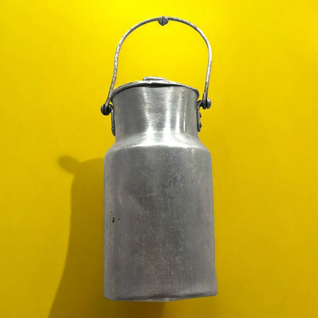 Lechera antigua de aluminio con tapa y asa metálica sobre fondo amarillo. Decoración vintage ideal para ambientes rústicos y coleccionistas.