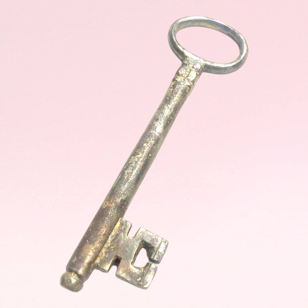Llave antigua de forja en metal envejecido sobre fondo rosa, ideal como elemento decorativo único para tu hogar en estilo vintage.
