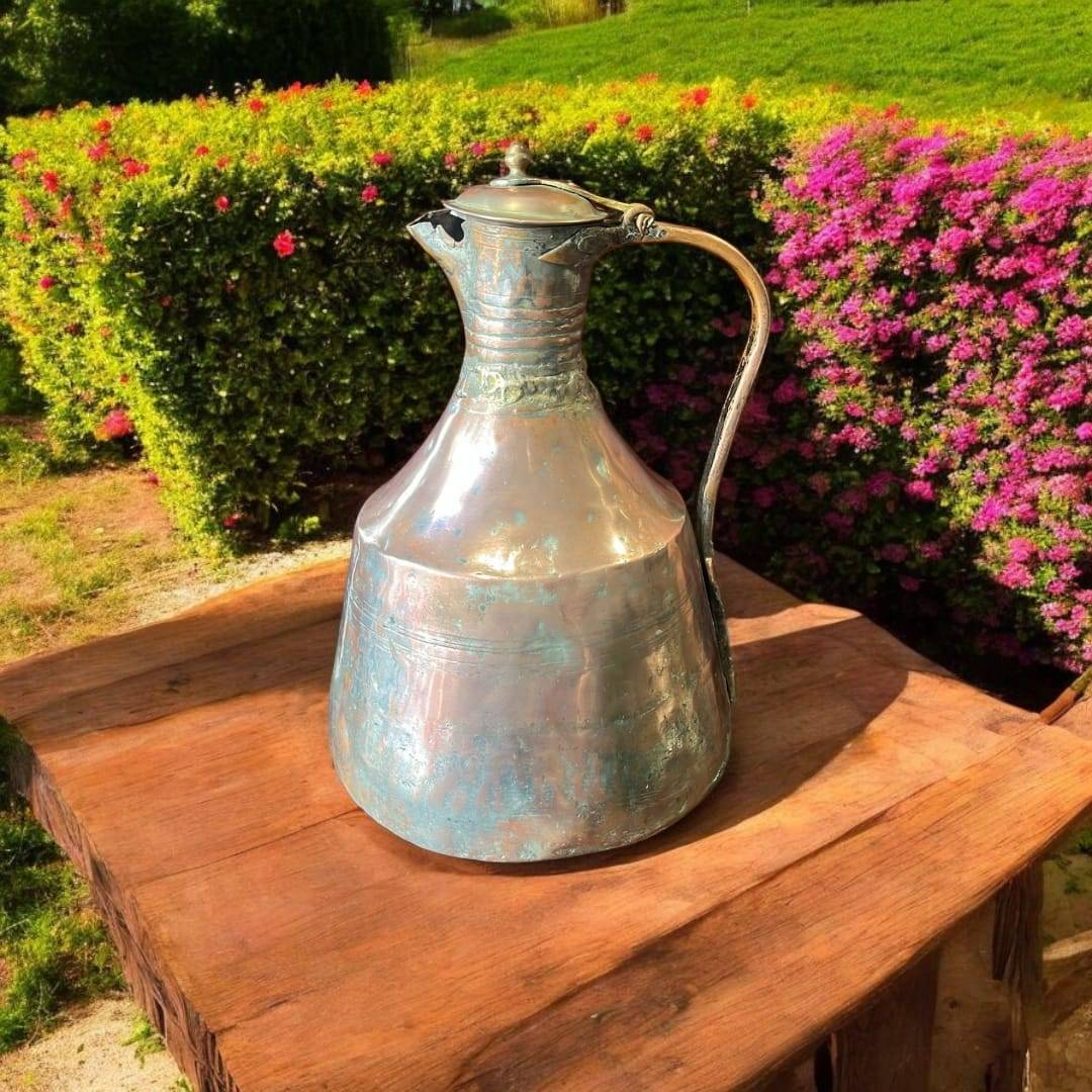 Jarra - Aguamanil antigua de cobre martillado artesanal con tapa y asa forjada, exhibida sobre mesa rústica en jardín con flores.