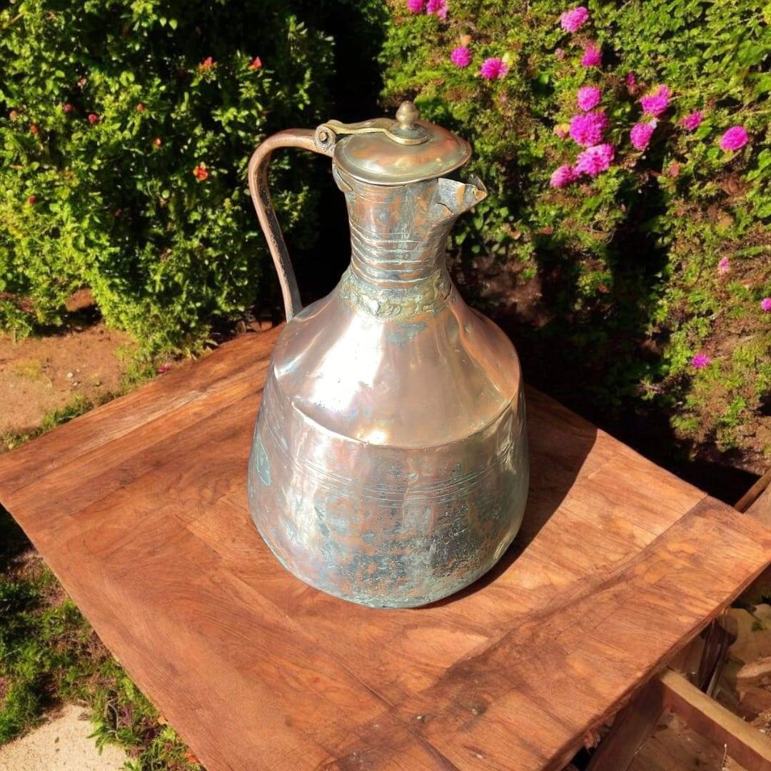 Jarra - Aguamanil antigua de cobre martillado artesanal con tapa y asa forjada, exhibida sobre mesa rústica en jardín con flores.