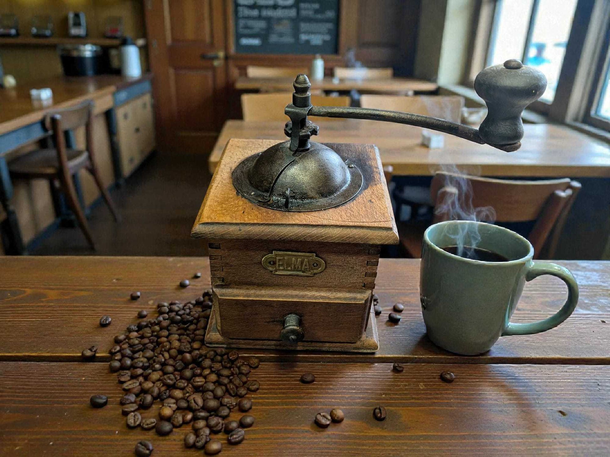 Molinillo de café antiguo Elma junto a taza humeante, sobre mesa rústica. Pieza única para decoración vintage en hogar o cafetería.