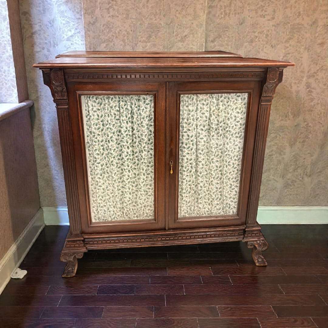 Vitrina de madera clásica con dos puertas y cortinas, ubicada junto a una ventana en un suelo de madera oscura. Ideal para decoración elegante.