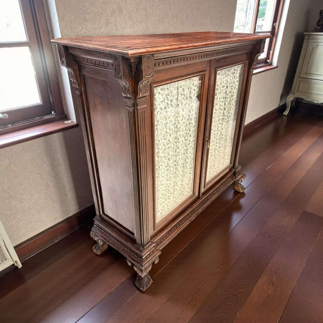 Vitrina de madera clásica con dos puertas y cortinas, ubicada junto a una ventana en un suelo de madera oscura. Ideal para decoración elegante.