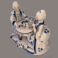 Figura decorativa de porcelana azul y blanca con pareja rococó tomando té, detalles florales y acabados dorados, ideal para decoración clásica.