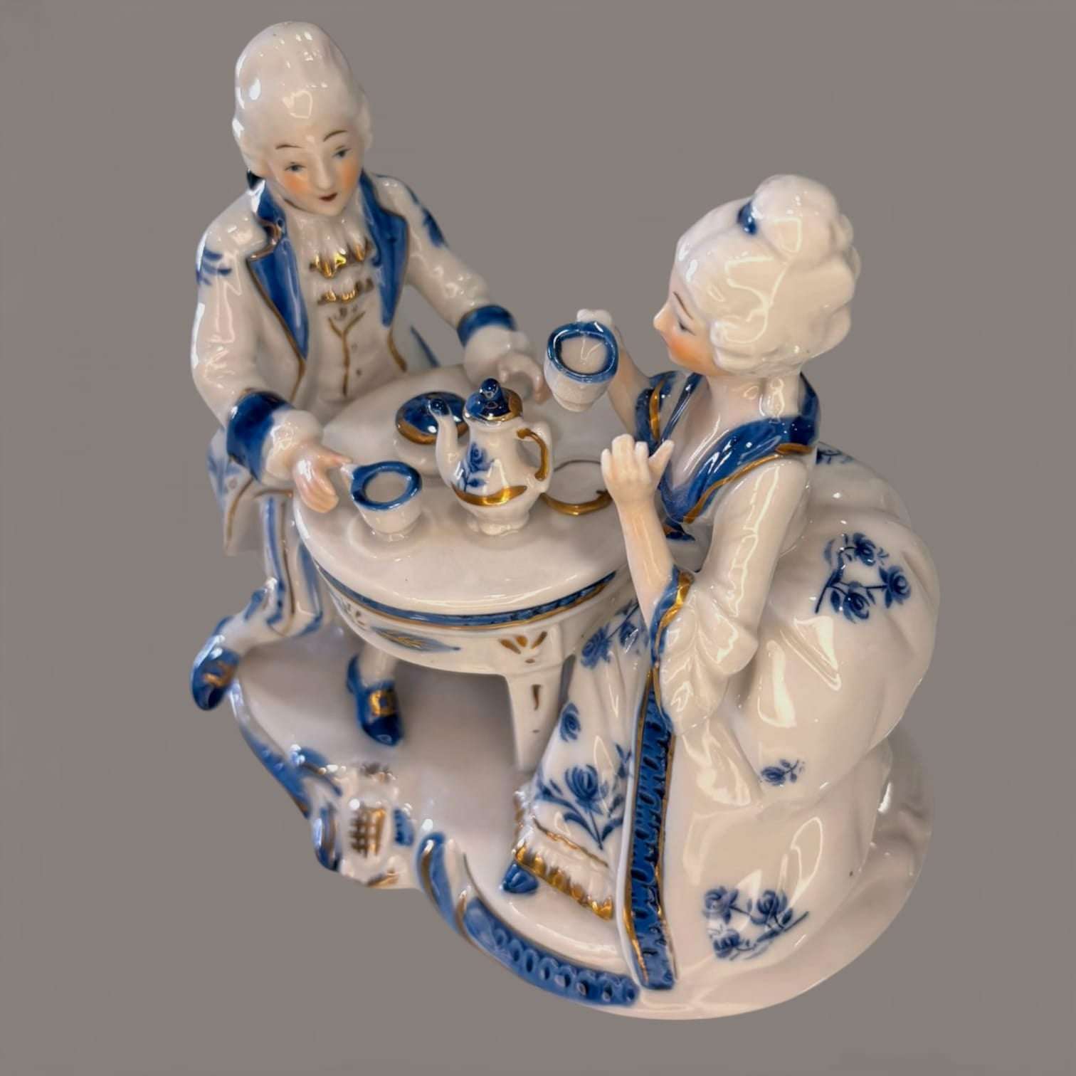 Figura decorativa de porcelana azul y blanca con pareja rococó tomando té, detalles florales y acabados dorados, ideal para decoración clásica.