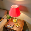 Lámpara sobre mesa metal pantalla burdeo claro 44 cm encendida, sobre mesa auxiliar junto a sofá beige y libros, ideal para interiores cálidos.