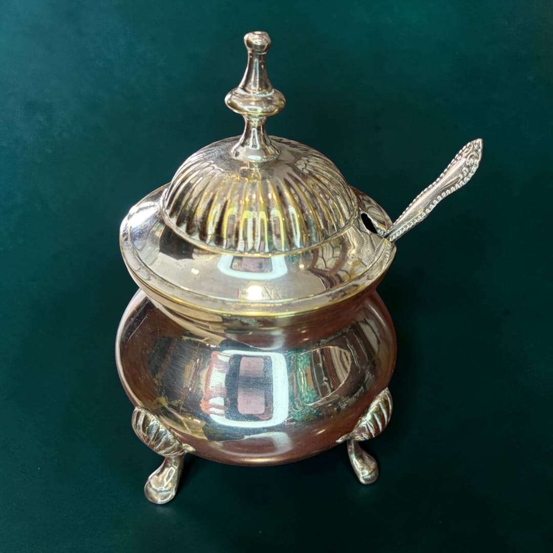 Este azucarero de metal plateado es una pieza de sobremesa concebida originalmente para guardar y servir azúcar de forma limpia, elegante y práctica durante el servicio de café, té o desayuno.