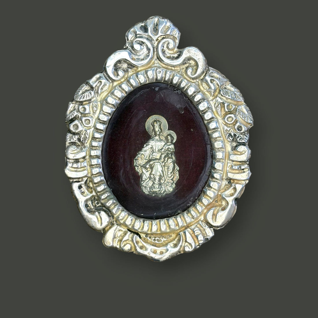 Este relicario de metal plateado con imagen de la Virgen del Carmen es una pieza devocional de pequeño formato, concebida originalmente para custodiar una imagen sagrada y acompañar la oración privada en el ámbito doméstico. 