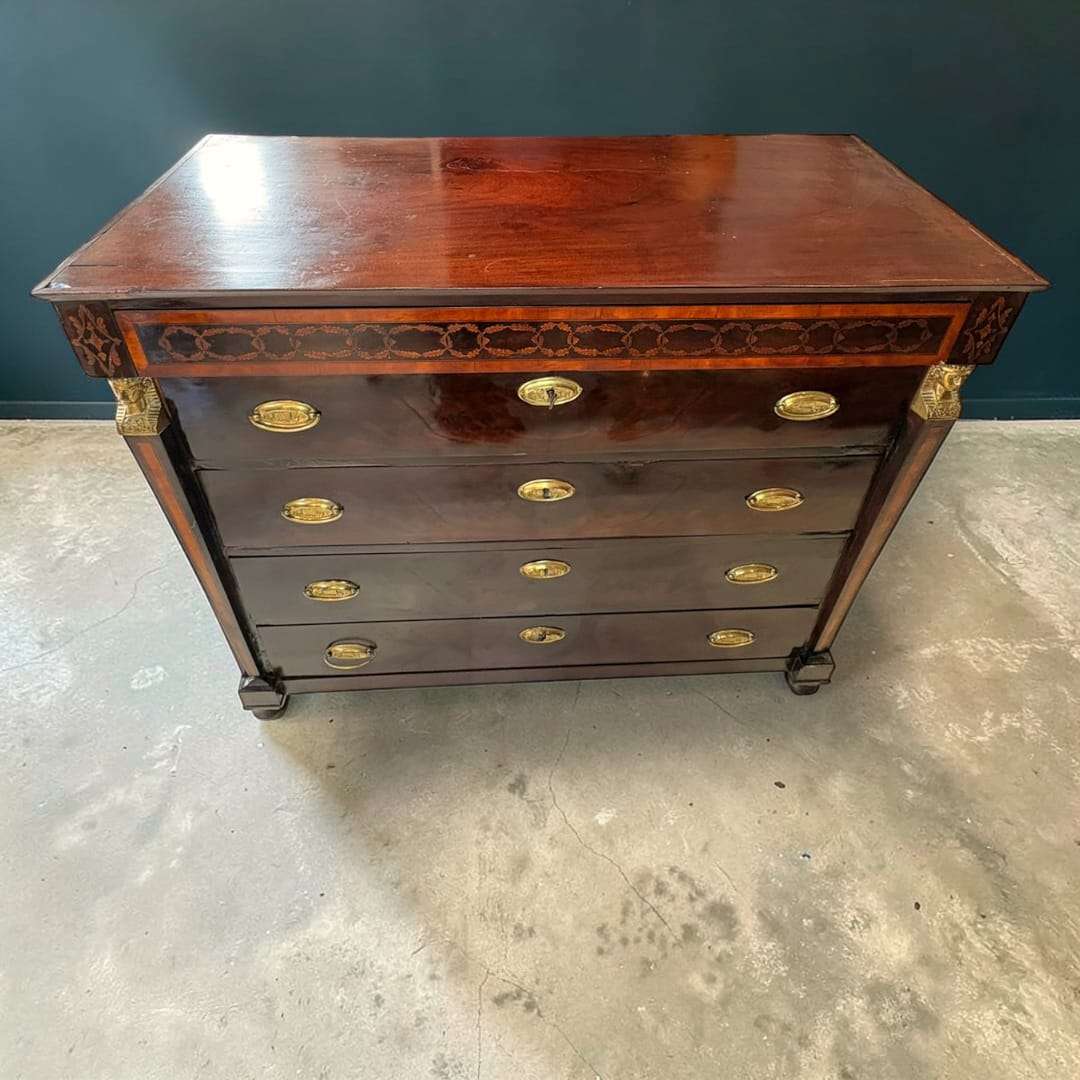 En las imágenes se aprecia una estructura sólida y refinada, con una silueta clásica realzada por sus columnas laterales características del estilo Imperio, su friso superior decorado con marquetería, y sus aplicaciones ornamentales metálicas en las esquinas superiores, que aportan un aire distinguido y señorial.