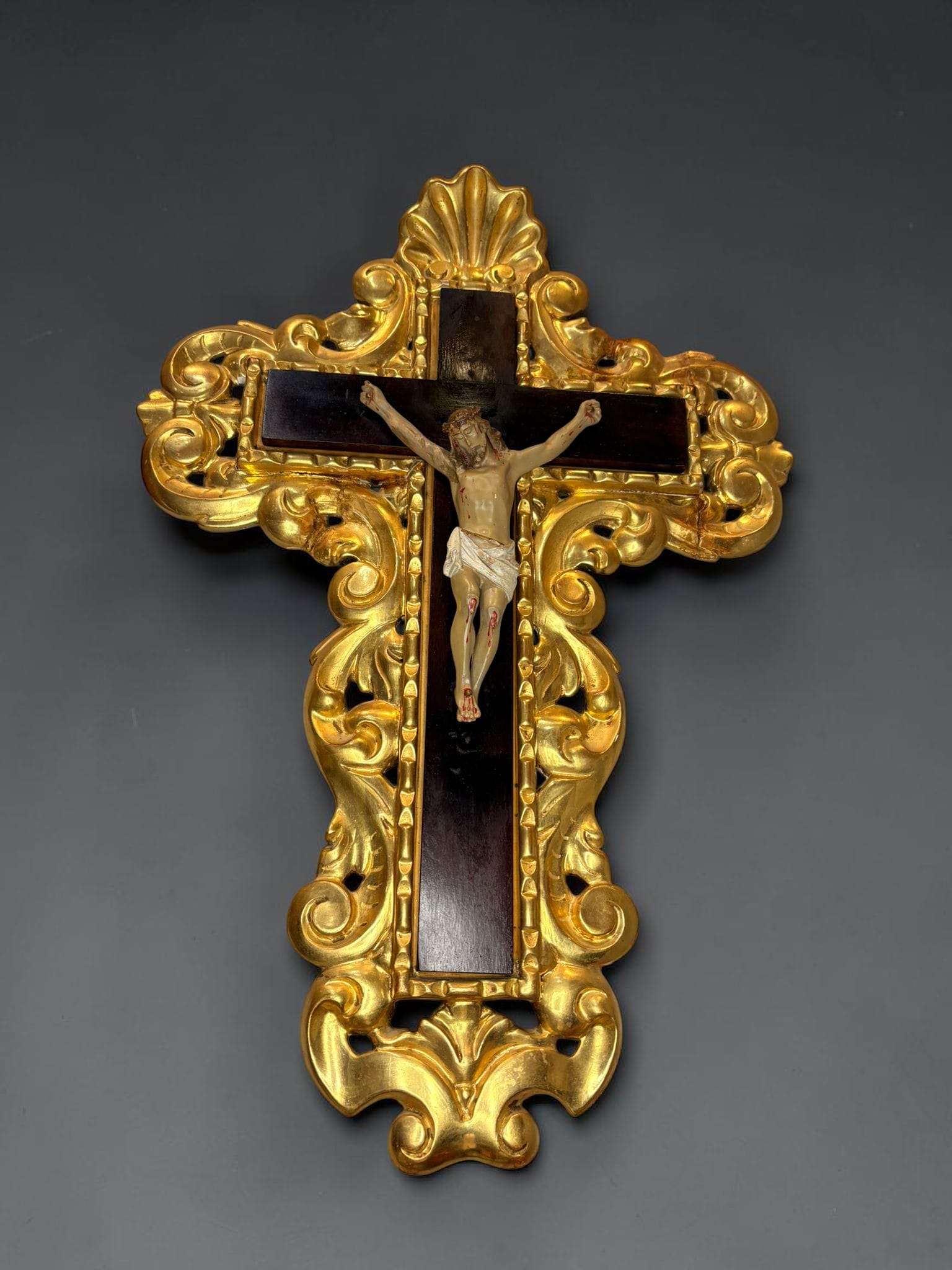 Se trata de una figura de Jesucristo crucificado tipo Olot, colocada sobre una cruz de caoba de tono oscuro y enmarcada por un espectacular contorno ornamental dorado tallado en madera. 