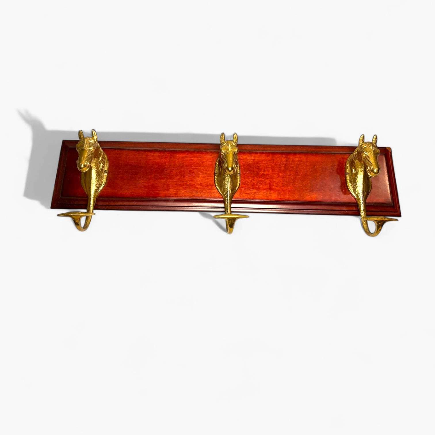 Realizado sobre una base de madera de tono caoba brillante, incorpora tres ganchos frontales con forma de cabezas de caballo en acabado bronce dorado, creando una composición visual refinada, equilibrada y con fuerte personalidad. 