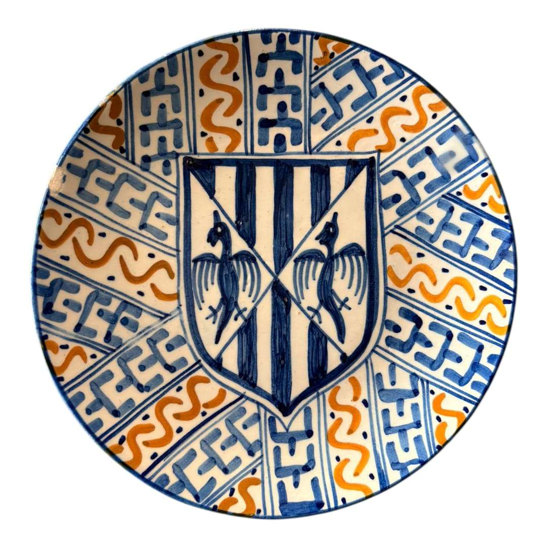 En las imágenes se aprecia una composición muy marcada, centrada en un escudo heráldico en azul sobre fondo claro, rodeado por una decoración radial de bandas geométricas y motivos ondulantes en azul cobalto y ocre anaranjado. 