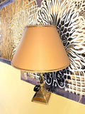 Presenta base cuadrada escalonada de latón, fuste central torneado con formas bulbosas, acabado dorado brillante, pantalla cónica beige con ribete superior e inferior, cable negro visible con interruptor de paso, formato compacto de sobremesa y una estética muy decorativa propia del gusto Hollywood Regency.