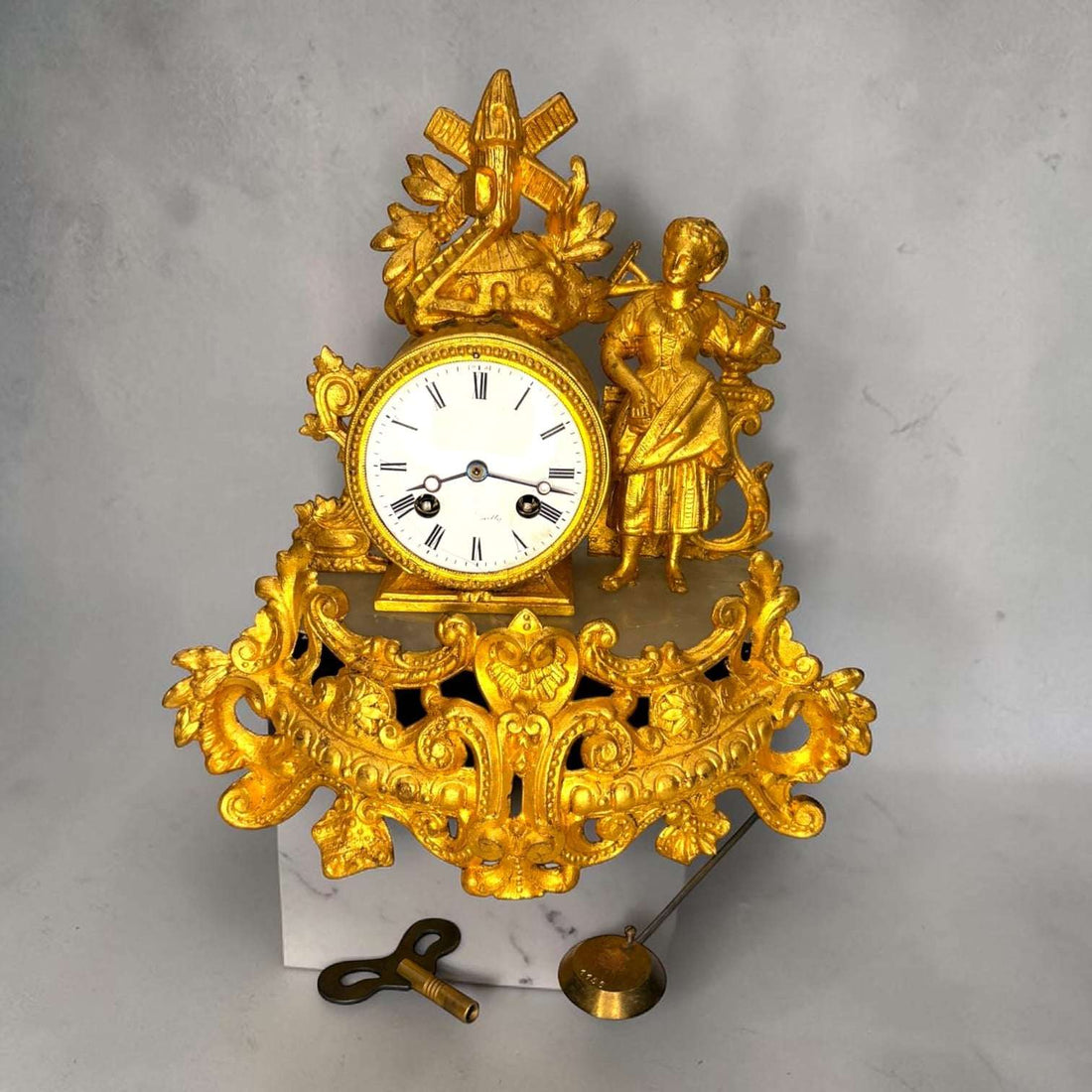 Este Reloj de sobremesa antiguo es una pieza decorativa de gran presencia, realizada en metal dorado y con una composición inspirada en el gusto rococó o Luis XV. 