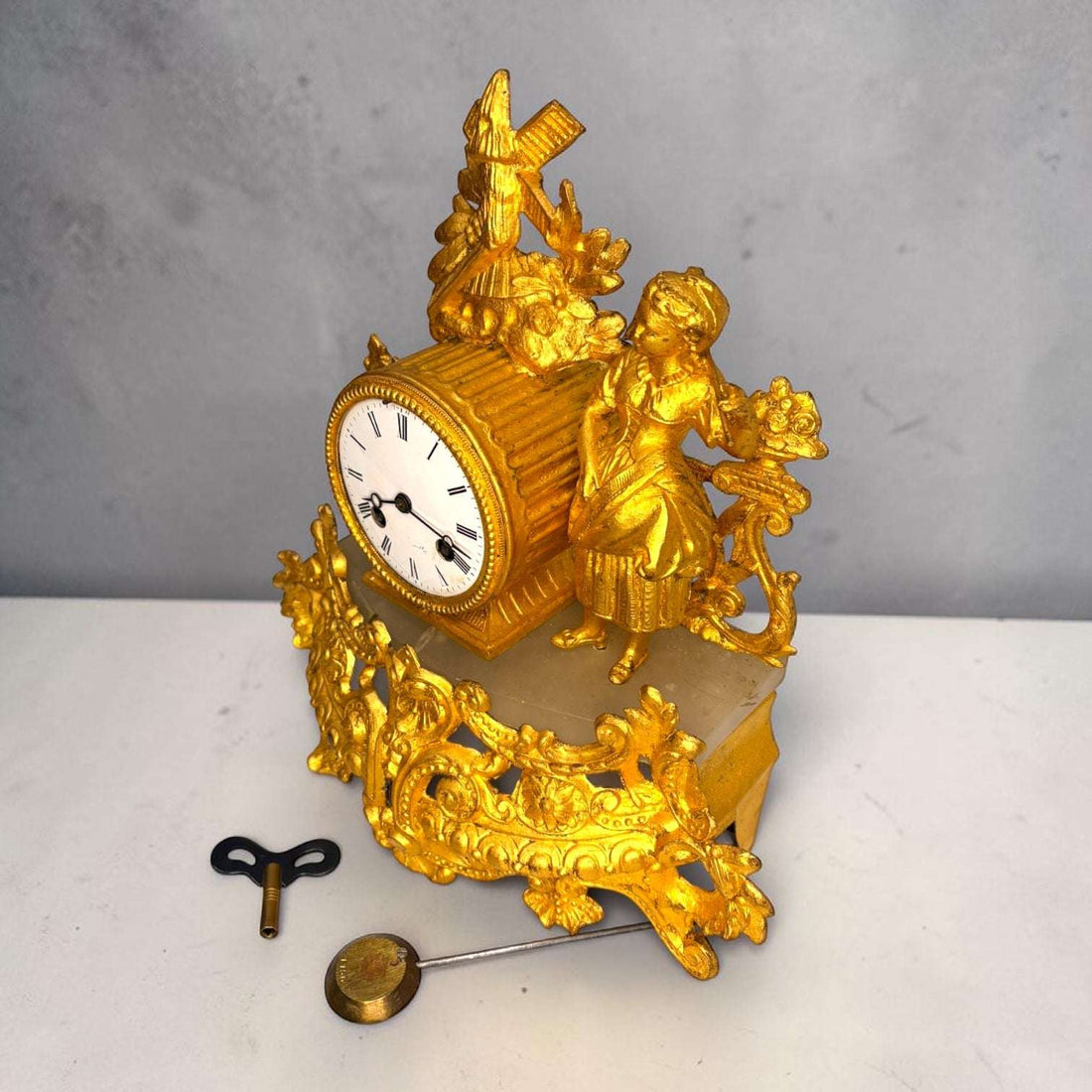 Este Reloj de sobremesa antiguo es una pieza decorativa de gran presencia, realizada en metal dorado y con una composición inspirada en el gusto rococó o Luis XV. 