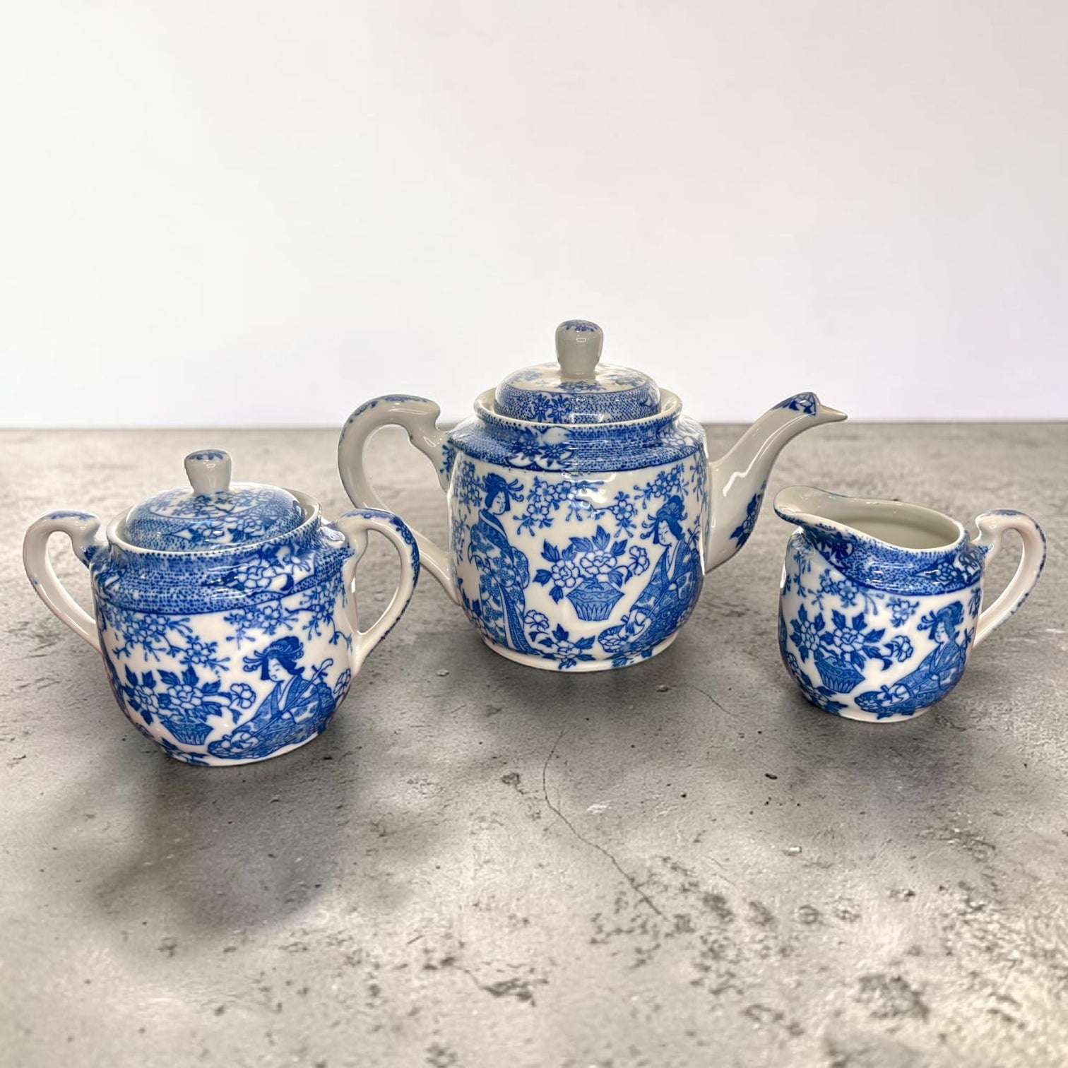 La cafetera presenta tapa, asa lateral y pico vertedor; el azucarero conserva tapa y doble asa; la lechera mantiene su forma pequeña con asa y boca de servicio; las tazas se acompañan de platos decorados a juego.