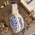 En la parte frontal se aprecia un motivo floral central de gran riqueza visual, rodeado de volutas, hojas y trazos en azul intenso sobre fondo blanco crema. El contraste entre ambos tonos aporta luminosidad, equilibrio y un aire clásico muy decorativo.