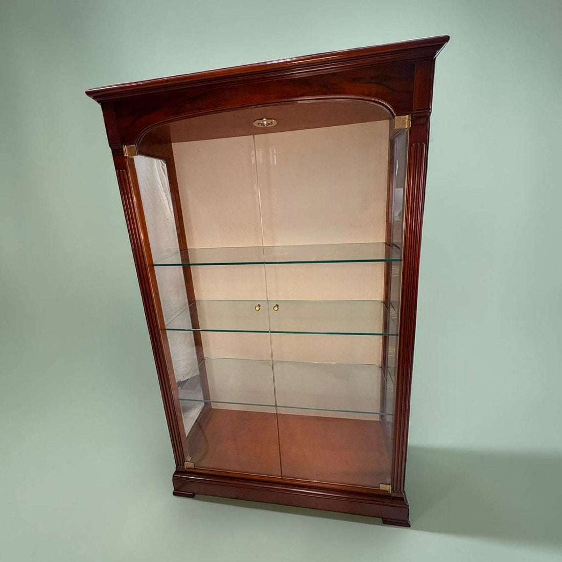 Esta Vitrina destaca por su diseño refinado, con una estructura alta y estilizada en madera de caoba de tono cálido y acabado brillante.