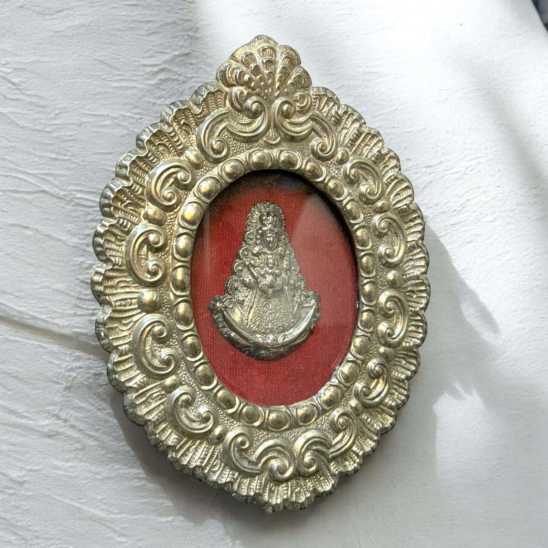 Este Relicario de metal plateado es una pieza devocional y decorativa de gran presencia, formada por un marco ovalado ornamentado y una imagen interior de la Virgen del Rocío sobre fondo textil rojo.