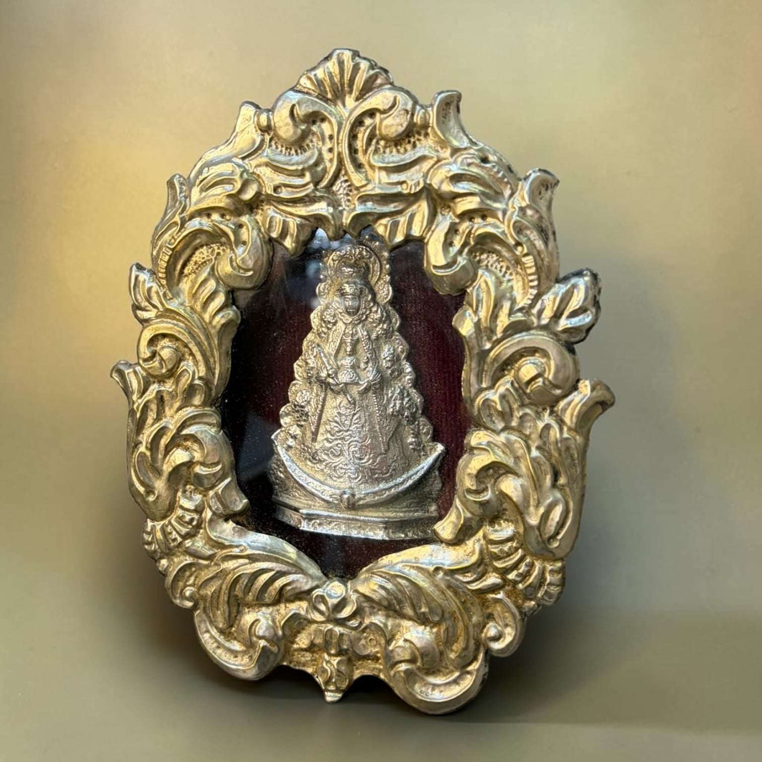 En su interior aparece una figura de la Virgen del Rocío en relieve metálico, situada sobre un fondo oscuro de tono granate, lo que crea un contraste sobrio, elegante y profundamente simbólico.