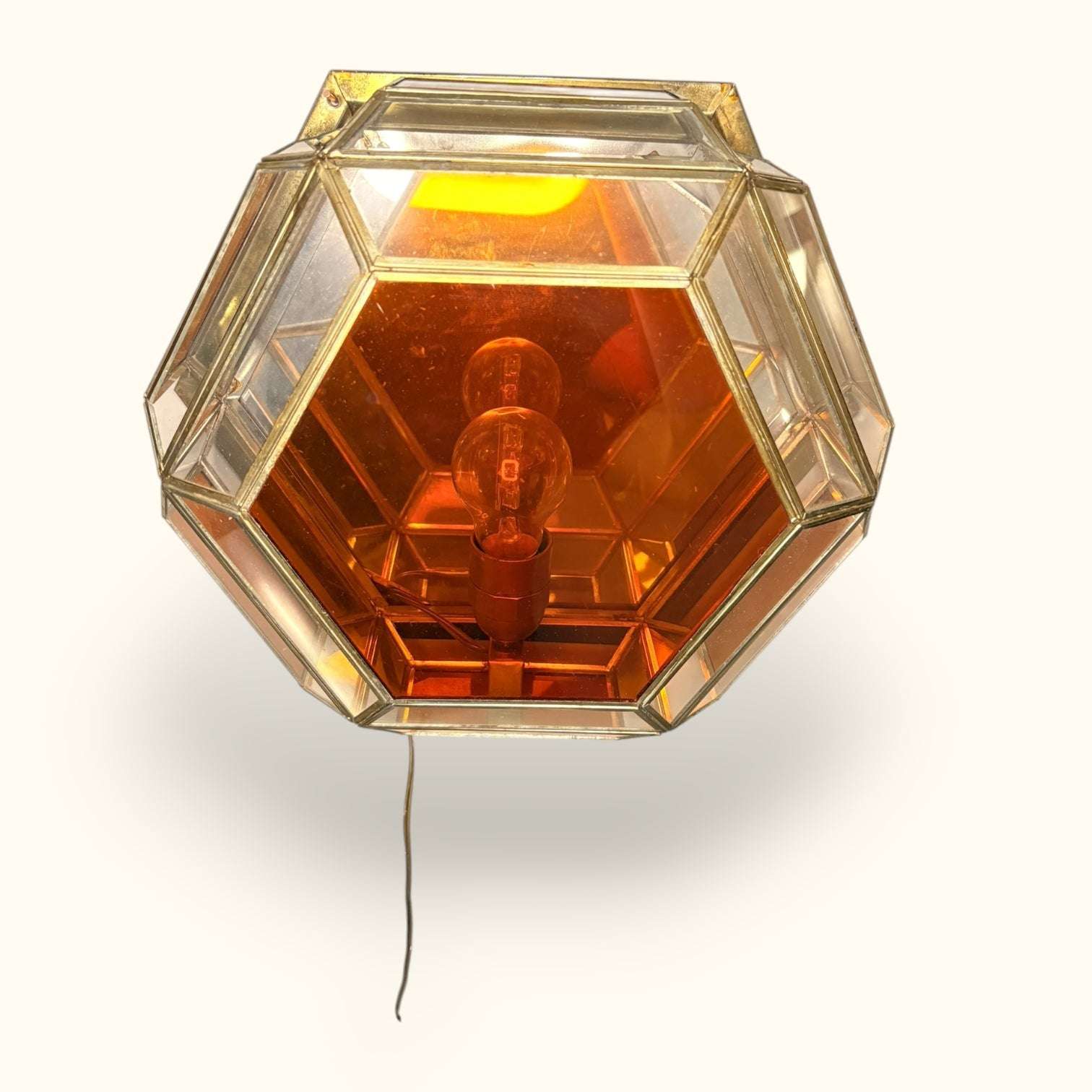 La Lámpara destaca por su composición geométrica: un cuerpo hexagonal con cristales biselados, perfiles metálicos dorados y una combinación de vidrio transparente con cristal color ámbar, que refuerza la sensación de calidez incluso cuando está apagada.