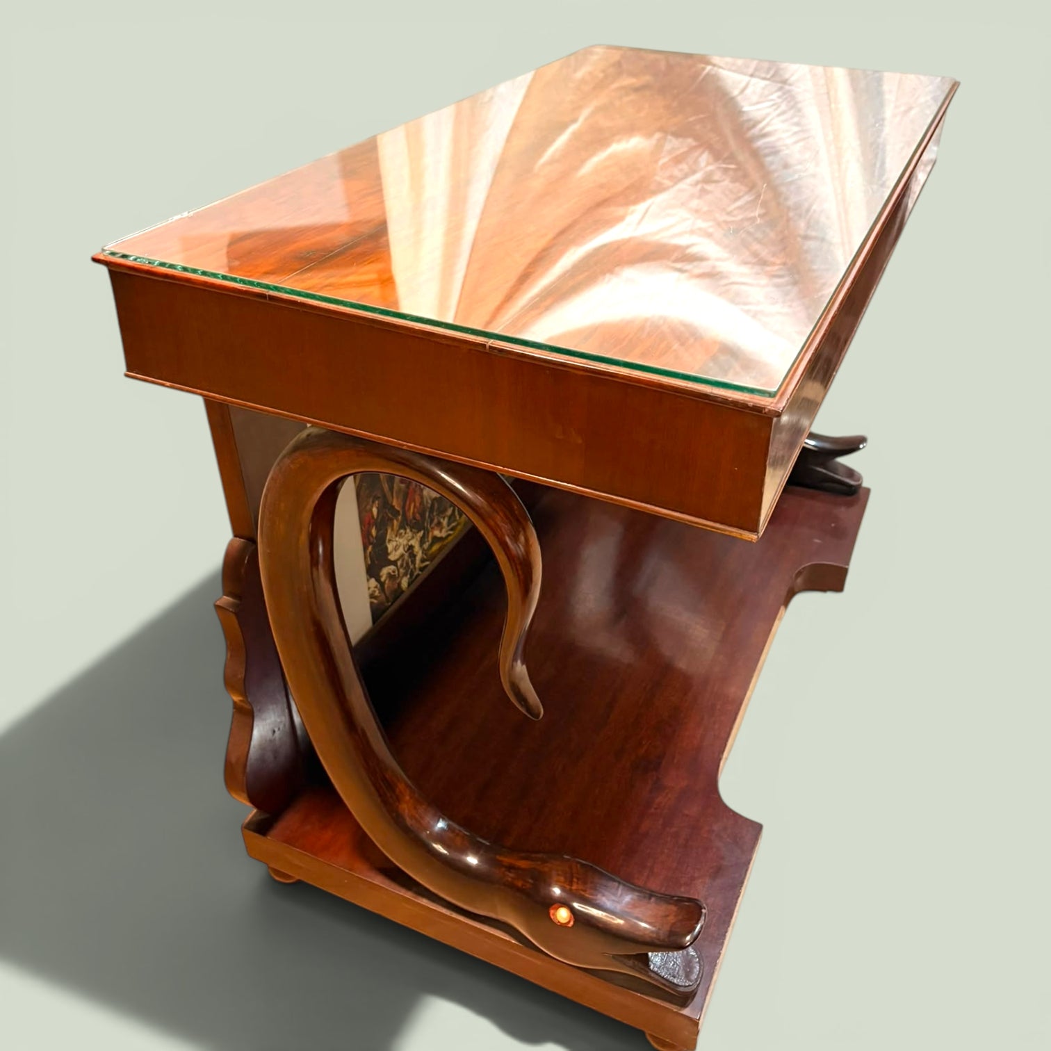 La consola de caoba con cristal destaca por su tapa superior con cristal protector, que aporta brillo y ayuda a conservar la superficie. La base inferior ofrece una repisa amplia, muy útil para decoración, mientras que los laterales curvos y los detalles tallados aportan movimiento, volumen y una estética muy reconocible.