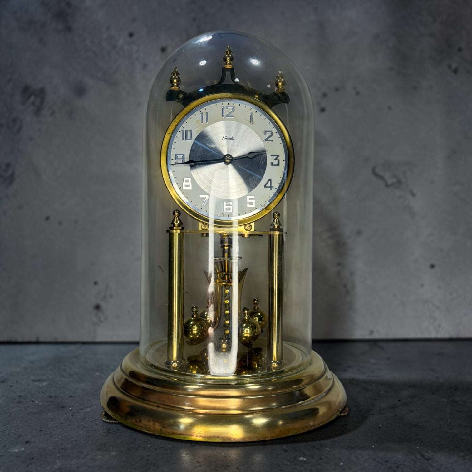 Reloj aniversario o reloj de péndulo de torsión marca Kundo, una pieza muy reconocible por su estructura de metal dorado, su mecanismo visible y su elegante cúpula de cristal.