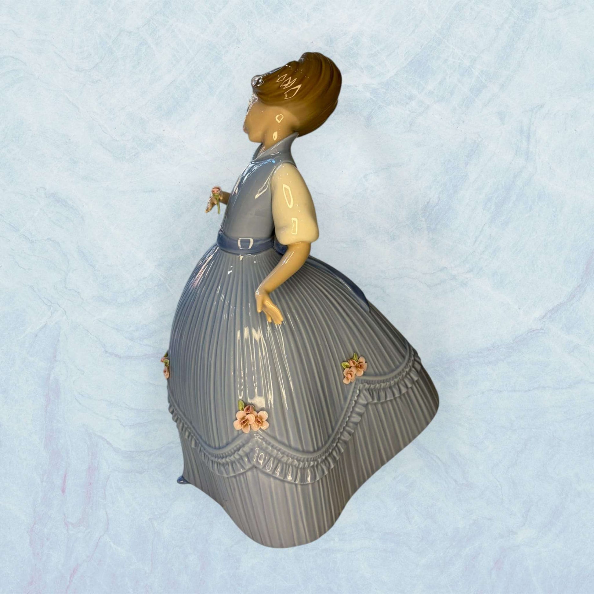 Su función principal es decorativa: embellecer vitrinas, aparadores, salones, dormitorios o espacios de colección donde se busque una pieza fina, luminosa y con presencia clásica.

La figura muestra a una niña de rostro sereno, con vestido azul de gran volumen, pliegues verticales muy marcados 