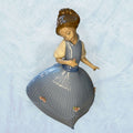 Su función principal es decorativa: embellecer vitrinas, aparadores, salones, dormitorios o espacios de colección donde se busque una pieza fina, luminosa y con presencia clásica.

La figura muestra a una niña de rostro sereno, con vestido azul de gran volumen, pliegues verticales muy marcados 
