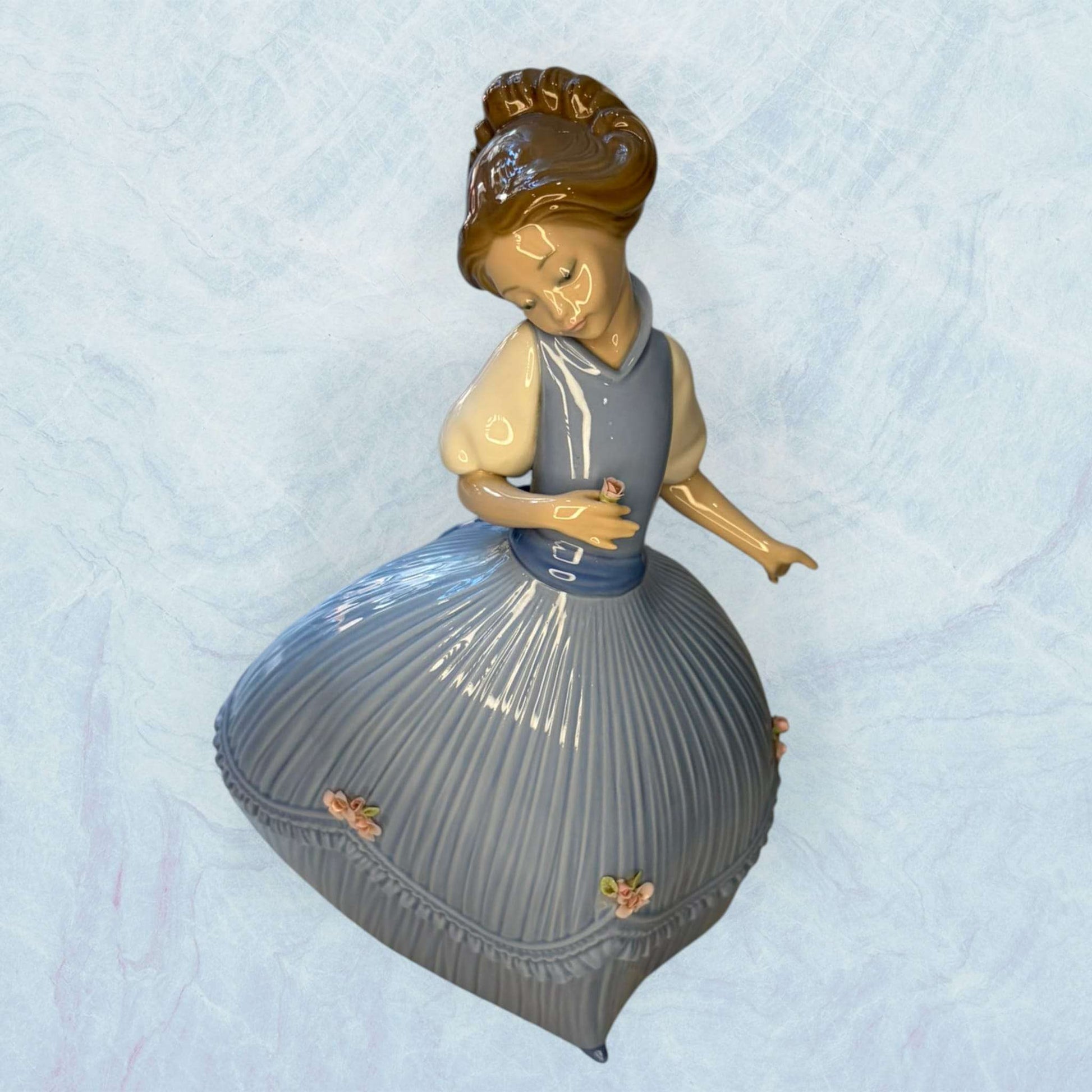 Su función principal es decorativa: embellecer vitrinas, aparadores, salones, dormitorios o espacios de colección donde se busque una pieza fina, luminosa y con presencia clásica.

La figura muestra a una niña de rostro sereno, con vestido azul de gran volumen, pliegues verticales muy marcados 