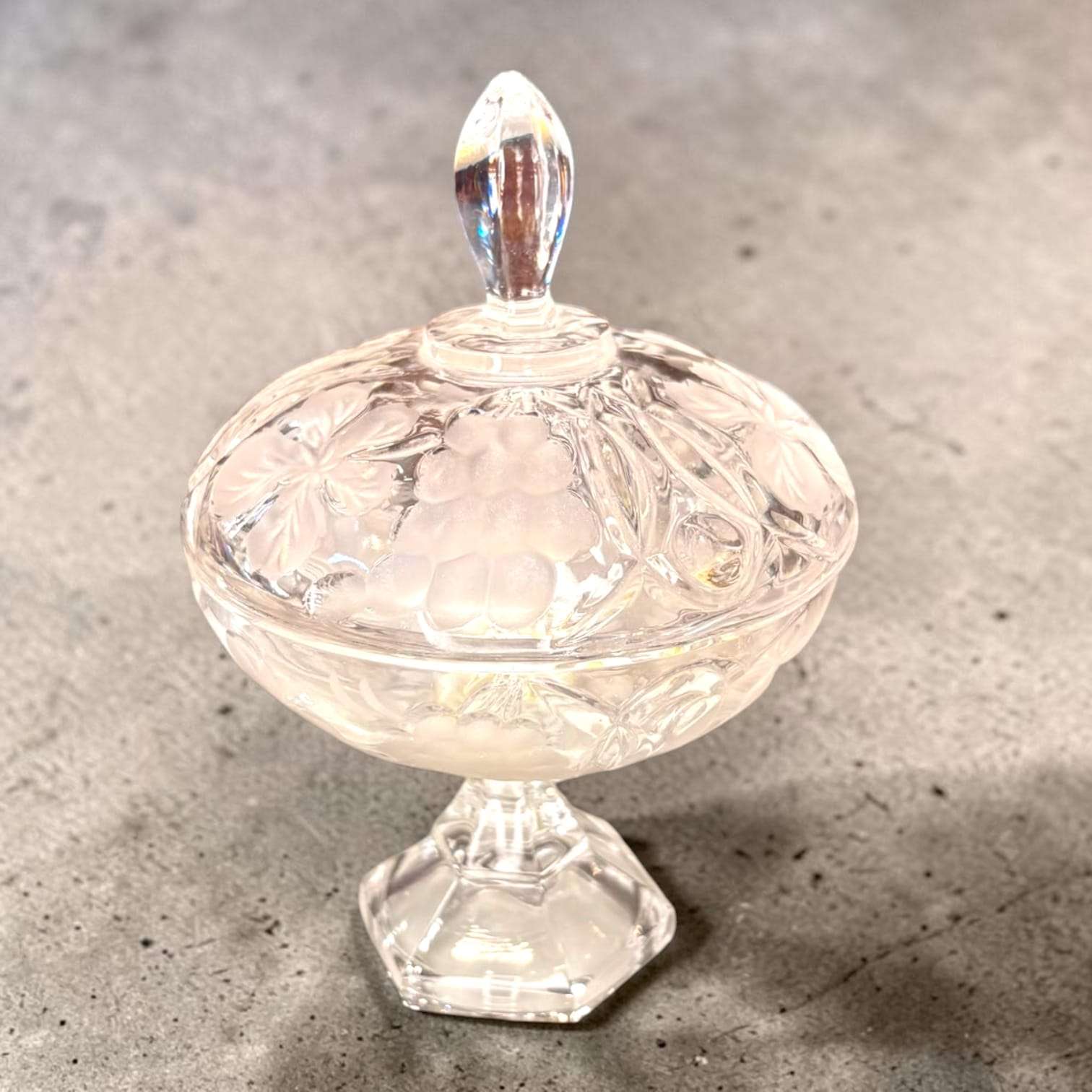 Vista superior de la bombonera de cristal con tapa, mostrando relieves florales y uvas, en acabado transparente y decorativo vintage.